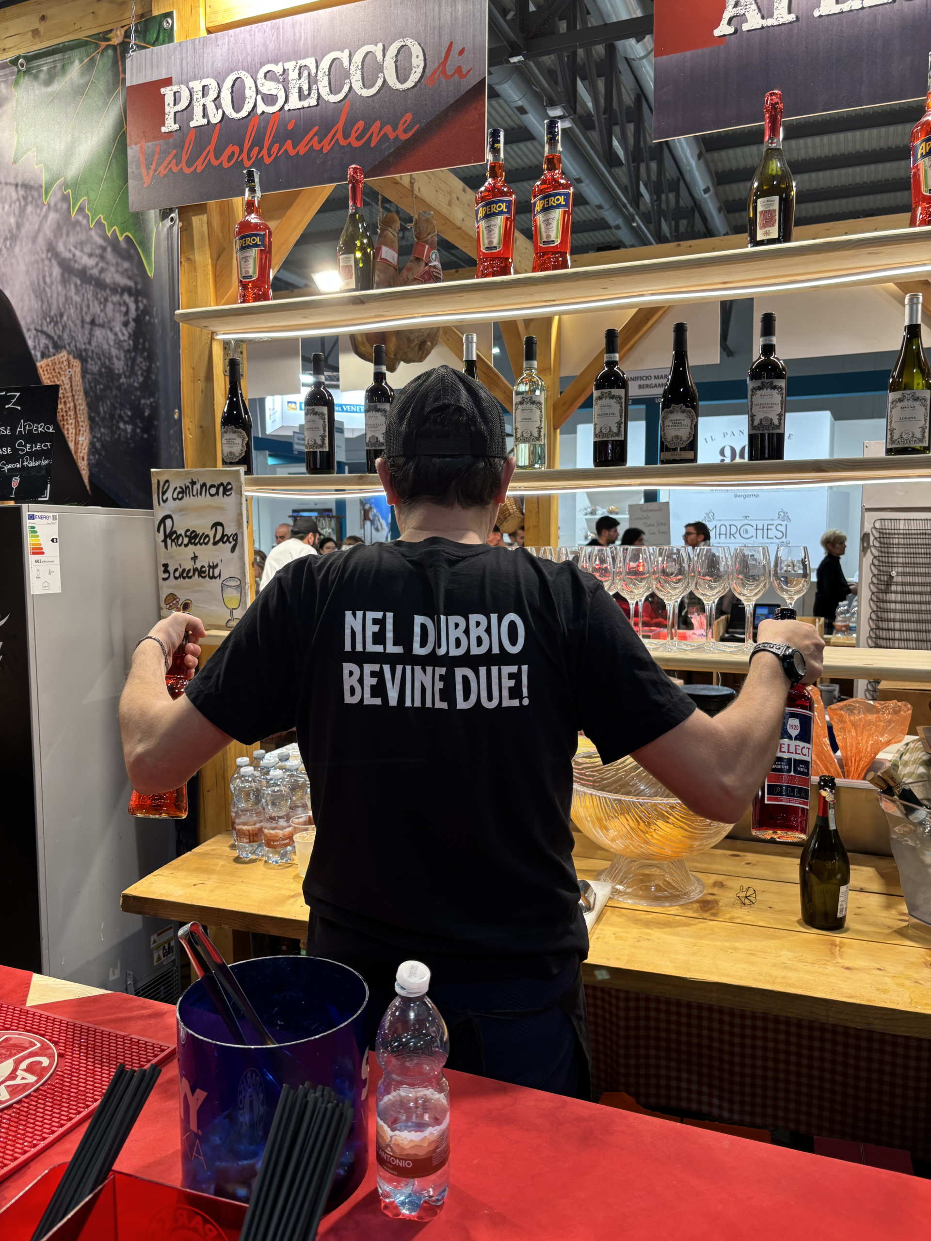 Barista con la maglietta «Nel dubbio bevine due!» al banco in fiera