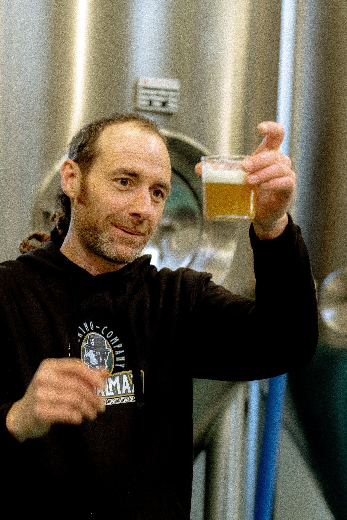 Mastro birraio brinda Mastro birraio alza un bicchiere di birra davanti ai tank